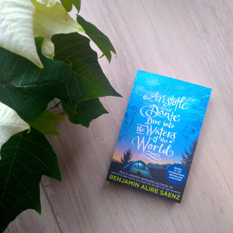 Bogen Aristotle and Dante Dive into the Waters of the World ligger på gråt trægulv ved siden af en hvid blomst med mørkegrønne blade