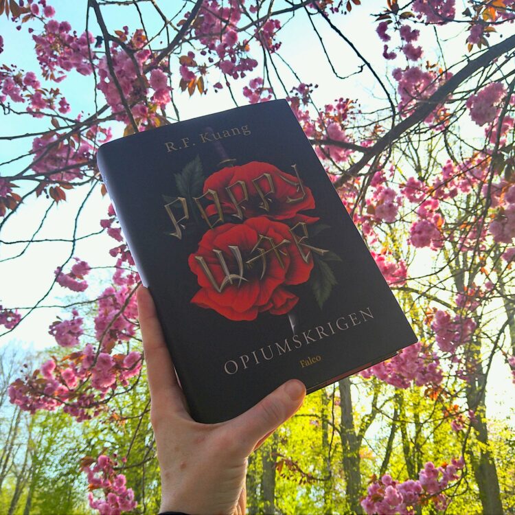 Bogen Poppy War Opiumskrigen holdes op imod en baggrund af lyserøde kirsebærblomster lysegrønne træer og en halvskyet blå himmel