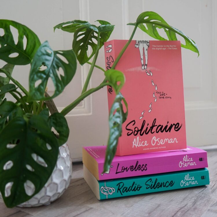 Bogen Solitaire står ovenpå andre bøger af Alice Oseman ved siden af en grøn plante